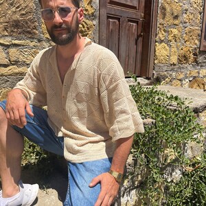 K&ouml;nnte beinhalten: Ein Mann tr&auml;gt ein beigefarbenes H&auml;kelhemd mit Knopfleiste und blaue Jeans-Shorts. Er tr&auml;gt eine goldene Uhr und eine Sonnenbrille.