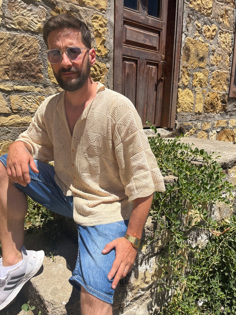 K&ouml;nnte beinhalten: Ein Mann tr&auml;gt ein beigefarbenes H&auml;kelhemd mit Knopfleiste und blaue Jeans-Shorts. Er tr&auml;gt eine goldene Uhr und eine Sonnenbrille.