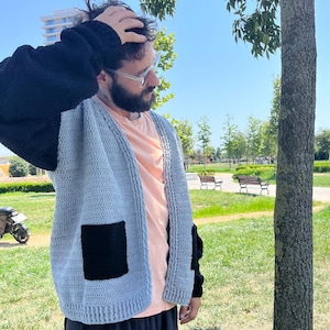 May include: A light blue knitted cardigan with black sleeves and a black patch pocket. The cardigan is open in the front and has a relaxed fit.