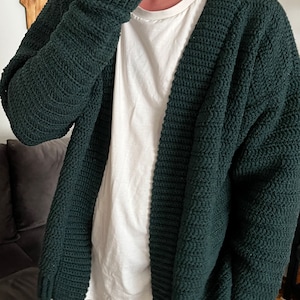 Może przedstawiać: Zielony, dzianinowy kardigan z głębokim dekoltem w kształcie litery V i długimi rękawami. Kardigan wykonany jest z miękkiej, fakturowanej przędzy i ma luźny krój.