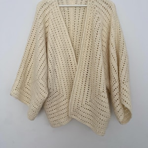 May include: A cream-colored crocheted cardigan with a loose fit and open front. The cardigan has a delicate, lacy pattern and long sleeves.