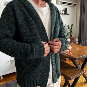 May include: A dark green knitted cardigan with a shawl collar and a button closure. The cardigan is made of a soft, textured yarn and has a relaxed fit.
