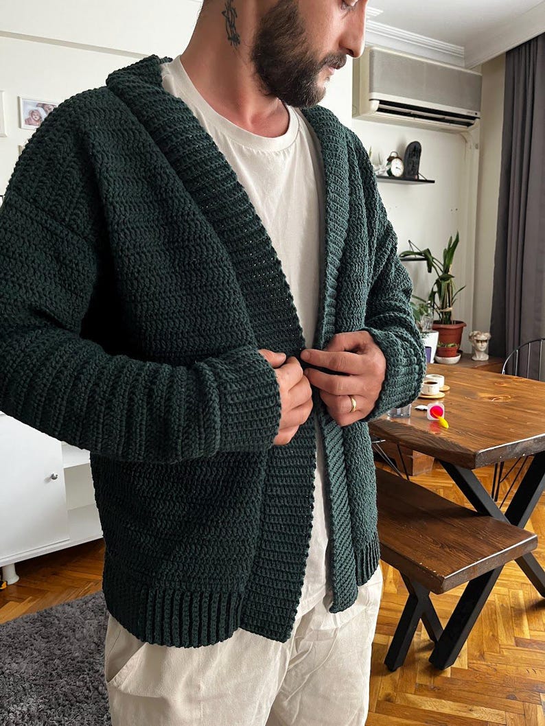 May include: A dark green knitted cardigan with a shawl collar and a button closure. The cardigan is made of a soft, textured yarn and has a relaxed fit.