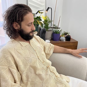 K&ouml;nnte beinhalten: Ein Mann in einem geh&auml;kelten, cremefarbenen Hemd sitzt auf einem wei&szlig;en Sofa. Er hat langes, dunkles Haar und einen Bart. Sein rechter Arm ist ausgestreckt und seine Hand ist offen.