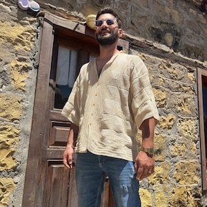 K&ouml;nnte beinhalten: Ein Mann tr&auml;gt ein beigefarbenes H&auml;kelhemd mit Kn&ouml;pfen, blaue Jeans-Shorts und eine Sonnenbrille. Er steht vor einem Steinhaus mit einer Holzt&uuml;r.