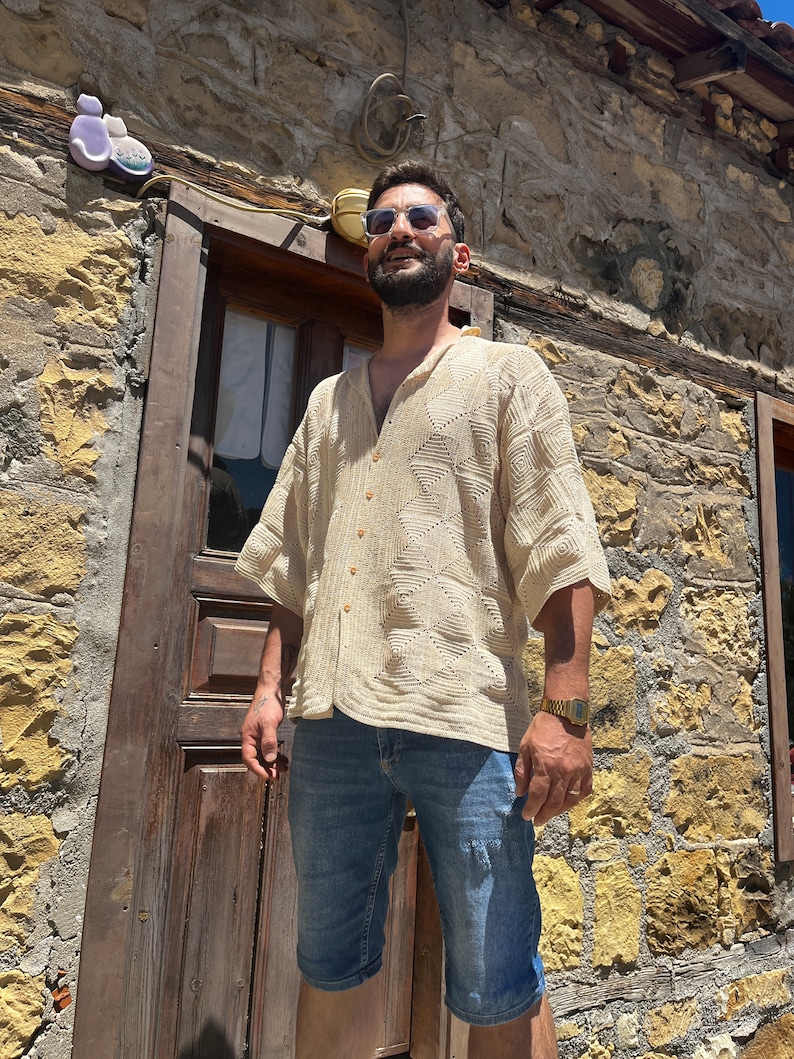 K&ouml;nnte beinhalten: Ein Mann tr&auml;gt ein beigefarbenes H&auml;kelhemd mit Kn&ouml;pfen, blaue Jeans-Shorts und eine Sonnenbrille. Er steht vor einem Steinhaus mit einer Holzt&uuml;r.
