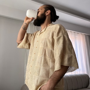 K&ouml;nnte beinhalten: Ein Mann tr&auml;gt ein beigefarbenes H&auml;kelhemd mit geometrischem Muster. Er h&auml;lt eine wei&szlig;e Tasse und hat einen Bart.
