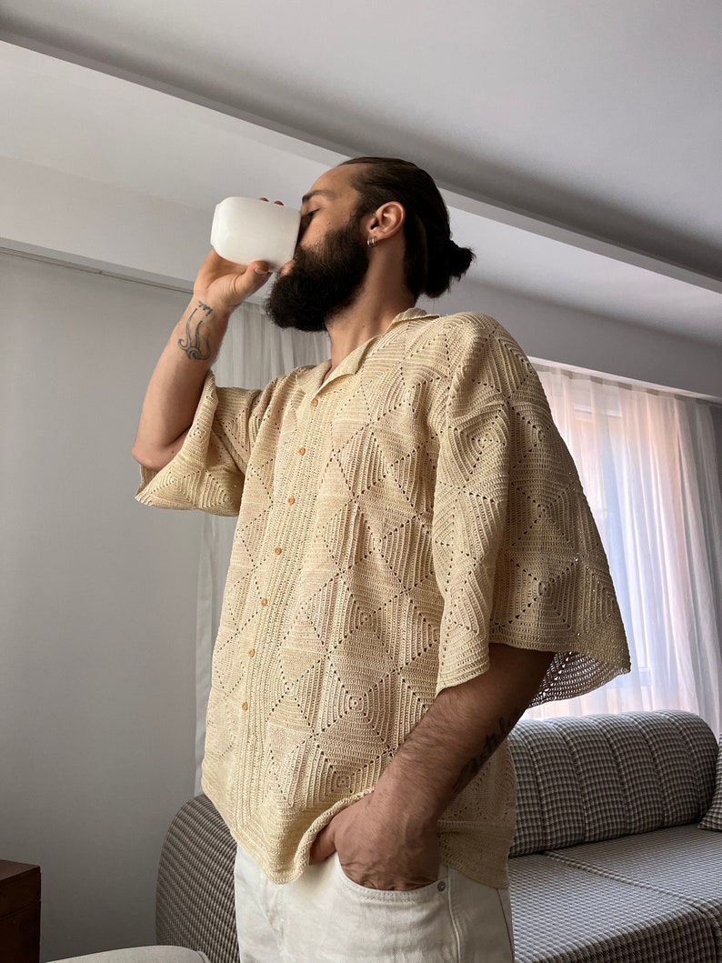 K&ouml;nnte beinhalten: Ein Mann tr&auml;gt ein beigefarbenes H&auml;kelhemd mit geometrischem Muster. Er h&auml;lt eine wei&szlig;e Tasse und hat einen Bart.