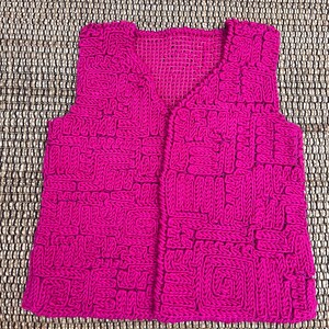 Peut inclure: Un gilet en tricot rose avec un motif géométrique. Le gilet a un col en V et est sans manches.