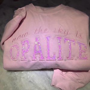 May include: A light pink sweatshirt with the words "now the sky is OPALITE" embroidered in a cursive font. The word "OPALITE" is in a block letter style and filled with sequins.