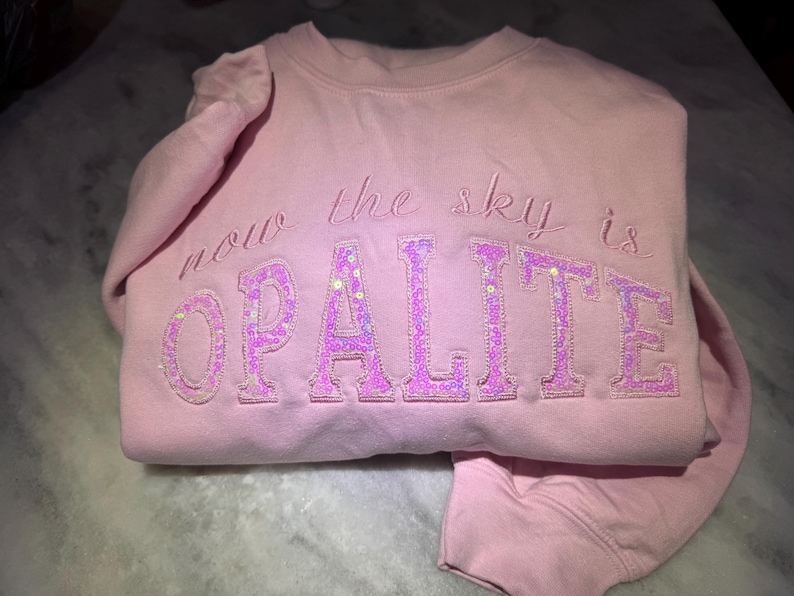 May include: A light pink sweatshirt with the words "now the sky is OPALITE" embroidered in a cursive font. The word "OPALITE" is in a block letter style and filled with sequins.