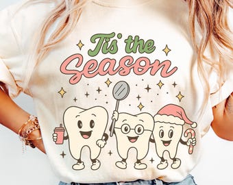 May include: An ivory t-shirt featuring the text 'Tis the Season' in green and pink script. The graphic includes three cartoon teeth characters: one holding a cup, one with glasses and a dental mirror, and one wearing a Santa hat holding a candy cane.