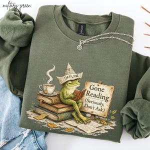 May include: Military green sweatshirt with a graphic of a frog reading a book, wearing a hat that says "OFFLINE". The sign reads "Gone Reading (Seriously, Don't Ask)". A cup of tea sits on a stack of books. A silver necklace is also visible.