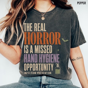 May include: A charcoal gray short-sleeve t-shirt featuring the text THE REAL HORROR IS A MISSED HAND HYGIENE OPPORTUNITY INFECTION PREVENTION in orange, purple, and cream colors, with a small green bat, a star, and a hand sanitizer bottle.