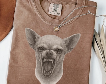 May include: Espresso-colored Comfort Colors t-shirt featuring a detailed illustration of an angry chihuahua with bared teeth. The shirt has a crew neck and short sleeves. A silver necklace with a small pendant is draped across the shirt.