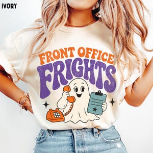 May include: An ivory t-shirt featuring the orange and purple text FRONT OFFICE FRIGHTS. The graphic includes a white ghost holding an orange rotary telephone and a blue clipboard with a star icon, accented by two small black four-pointed stars.