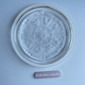 May include: A clear glass plate with a white rim and a circular design etched into the surface. The design features a sun with rays, a skull, a bird, a flower, and other symbols. The text "SAMHAIN", "YULE", "IMBOLC", "OSTARA", "BELTANE", "LITHRA", "MIDSUMMER", "LAMMAS" is etched around the edge of the plate.