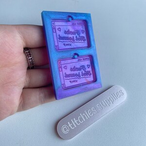 May include: Two clear purple and blue resin molds with a glittery finish. The molds are shaped like a computer screen and have the text "Get Journal" and "Sweet" inside a heart. The molds are sitting on a white surface.