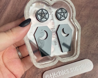 May include: A clear resin mold with two pentagram earrings and two coffin-shaped earrings with crescent moons. The text "MoonChild 1983" and "titchies.supplies" are also visible.