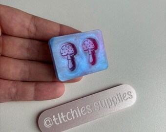 May include: A clear resin mold with two mushroom designs. The mold is a light blue color with a glittery finish. The mushrooms are a light purple color with white dots.