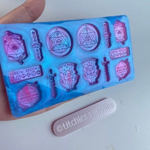 May include: A clear blue and pink resin mold with various shapes and designs. The mold features a variety of geometric shapes, including circles, triangles, and squares. The mold also includes a sword, a dice, and a hand holding a dice. The mold is perfect for creating unique and personalized jewelry, keychains, or other crafts.