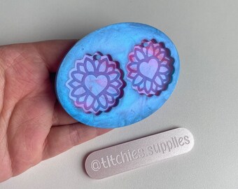 May include: A blue and white resin mold with two heart-shaped designs inside. The designs are outlined in white and have a pink and white marbled effect. The mold is oval shaped and has a textured surface. The text "@titchies.supplies" is printed on a silver rectangle.
