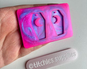 May include: Pink and blue resin mold with two coffin-shaped earring cavities. Each cavity has a crescent moon cutout. The mold is on a hand with a silver sticker that says "@titchies.supplies".