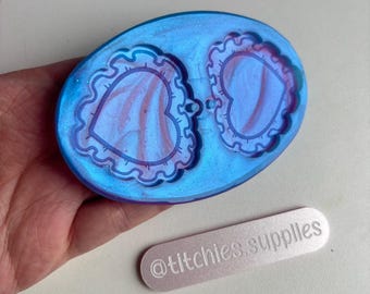 May include: A blue and purple resin mold with two heart-shaped cavities. The hearts have a scalloped edge and a swirl design. The mold is on a hand holding a white rectangle with the text "@titchies.supplies".