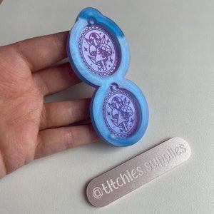 Peut inclure: Un moule en résine bleu et violet avec deux cavités circulaires. Chaque cavité présente un motif de deux champignons avec des étoiles et une lune croissante. Le moule est destiné à la fabrication d'objets en résine. Le moule est sur une main et un rectangle blanc avec le texte "@titchies.supplies" est au premier plan.