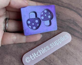 May include: A purple glitter resin mold with two mushroom shapes. The mold is in the shape of a rectangle and has a white background. The mushrooms are outlined in white and have white polka dots. The mold is being held by a hand. A white rectangle with the text "@titchies.supplies" is in the background.