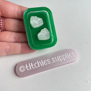 Könnte beinhalten: Zwei kleine weiße Kuchen-Anhänger in einer grünen Harzschale. Die Schale liegt auf einer rechteckigen Oberfläche in Rosa und Weiß mit dem aufgedruckten Text "@titchies.supplies".