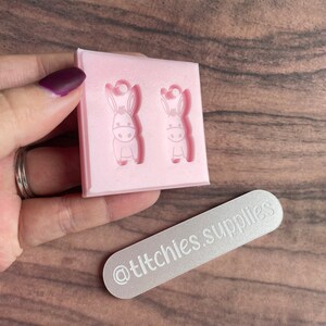 May include: Pink silicone mold with two donkey shaped cavities. The mold is being held by a hand. A silver rectangle with the text "@titchies.supplies" is in the background.