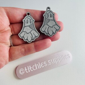 May include: Two grey wooden ghost ornaments with stitched details and a black outline. The ornaments have a hole at the top for hanging. The ornaments are on a hand with a pink and white background. The background also has the text "@titchies.supplies".
