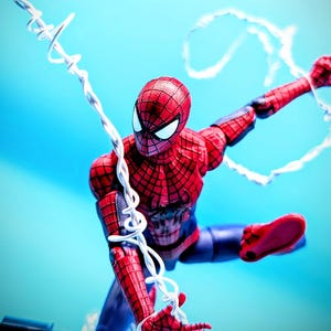 May include: A red and blue Spider-Man action figure in a dynamic pose, featuring a textured suit with a black web pattern and large white eyes. The figure is suspended in mid-air, holding white plastic web strands that extend across the frame.