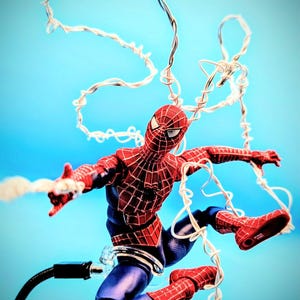 May include: A red and blue Spider-Man action figure in a dynamic pose, featuring a black web pattern across the red sections of the suit. The figure is surrounded by white, string-like web accessories that loop around the body and limbs.