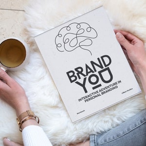 May include: A person is holding a workbook titled "Brand You: Interactive Adventure in Personal Branding" with a brain illustration on the cover. The workbook is on a white fluffy surface.