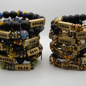 Könnte beinhalten: Zwei Stapel Armbänder mit Perlen. Die Armbänder haben schwarze matte Perlen, goldfarbene Akzente und rechteckige Namensschilder. Einige Perlen haben bunte Muster. Die Namensschilder tragen Namen wie "Kwaku" und "Afua".