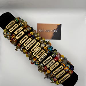 May include: A collection of beaded bracelets featuring rectangular gold-toned beads with black lettering. Each bead displays a unique name including Esther, Heaven, Afia, Adon, Maane, Julie, Blessing, Miracle, Evelyn, Daavi, Selasi, Serwaa, Rose, Adwoa, Kwabena, Junior, and Nyamekye.