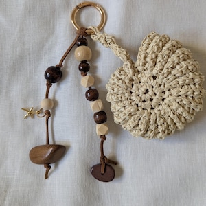 Pode incluir: Um chaveiro artesanal com um coração de ráfia tecido, contas de madeira e um fecho dourado. O chaveiro inclui contas de madeira castanho escuro, castanho claro e natural, e um pequeno berloque dourado. O coração de ráfia é bege.