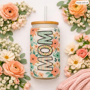 May include: A glass tumbler with a bamboo lid and straw, featuring a floral pattern in shades of peach, pink, and teal. The word MOM is printed vertically in large, block letters with a plaid and striped pattern on the front of the glass.