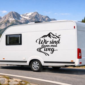May include: A white travel trailer featuring a black vinyl decal on its side. The decal displays a graphic of a mountain range with birds above it, a winding road below, and the German text "Wir sind dann mal weg" in a script font.