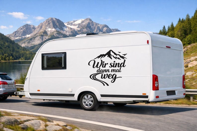 May include: A white travel trailer featuring a black vinyl decal on its side. The decal displays a graphic of a mountain range with birds above it, a winding road below, and the German text "Wir sind dann mal weg" in a script font.