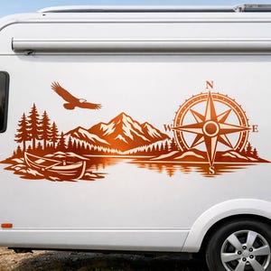 May include: A copper-colored vinyl decal applied to the side of a white camper van. The graphic features a mountain range, a forest of pine trees, a small boat on water, a flying eagle, and a large compass rose with the letters N, S, E, and W.