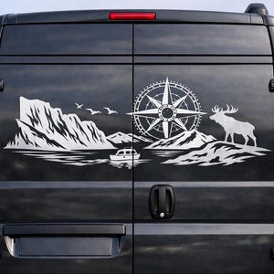 Puede incluir: Una furgoneta negra presenta un gran adhesivo blanco en sus puertas traseras. El gr&aacute;fico muestra un paisaje monta&ntilde;oso con una rosa de los vientos, un peque&ntilde;o barco en el agua, tres aves volando y la silueta de un alce de pie.