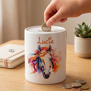 May include: A white ceramic piggy bank with a colorful watercolor horse design and the name "Lucia" in orange script. A hand is inserting a coin into the slot. Several coins are on the wooden surface.