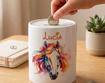 Puede incluir: Una hucha de cerámica blanca con un diseño de caballo en acuarela colorida y el nombre "Lucia" en escritura naranja. Una mano introduce una moneda en la ranura. Varias monedas están sobre la superficie de madera.