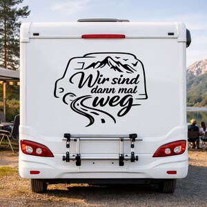 May include: A white camper van featuring a black vinyl decal on the rear door. The decal displays a stylized mountain range, a winding road, and the German text Wir sind dann mal weg, which translates to We are off now, positioned above a bike rack.