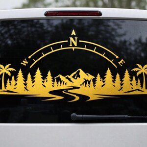 May include: A gold-coloured vinyl decal applied to a vehicle window, featuring a landscape silhouette of palm trees, pine trees, and mountains with a winding river in the foreground, topped by a compass arc marked with the letters N, W, and E.