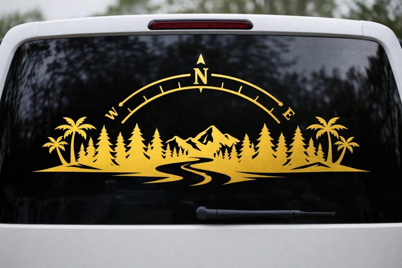 May include: A gold-coloured vinyl decal applied to a vehicle window, featuring a landscape silhouette of palm trees, pine trees, and mountains with a winding river in the foreground, topped by a compass arc marked with the letters N, W, and E.