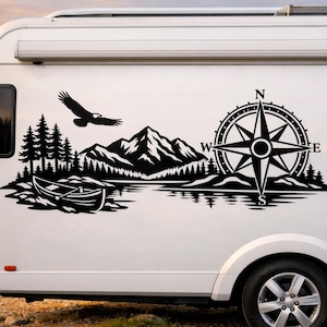 May include: A black vinyl decal applied to the side of a white recreational vehicle. The graphic features a mountain range, a forest of pine trees, a canoe on water, a flying eagle, and a large compass rose with the letters N, S, E, and W.